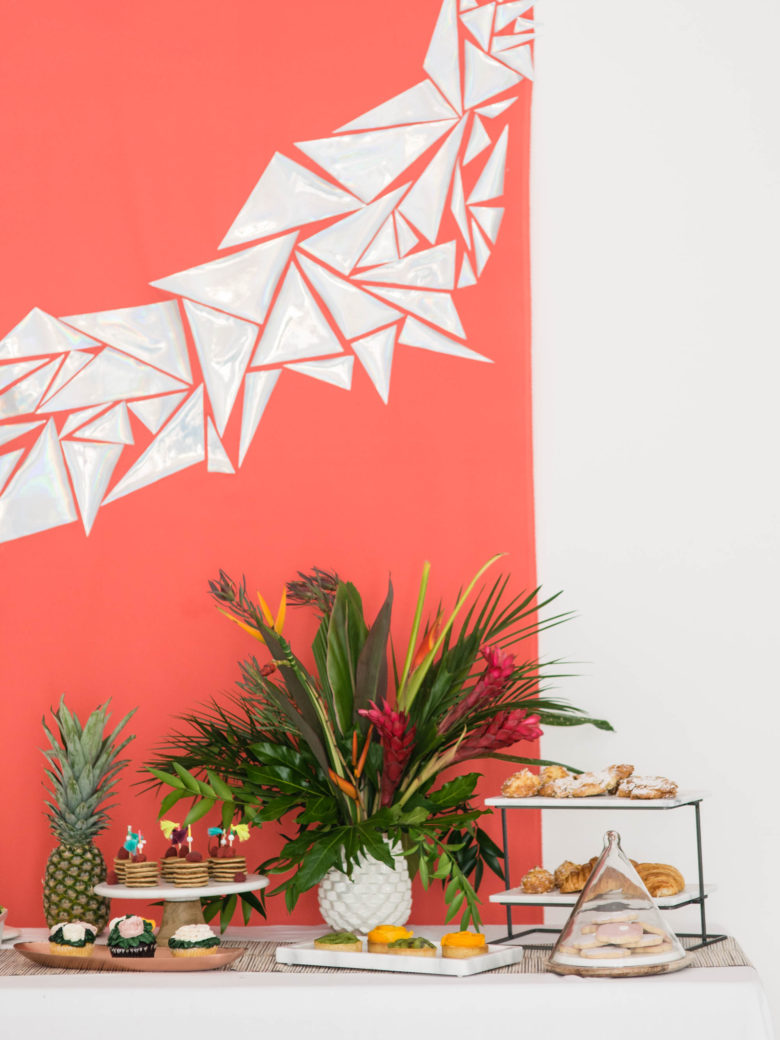 brunch party spread in front of coral backdrop with holographic triangles, with tropical plants and a pineapple