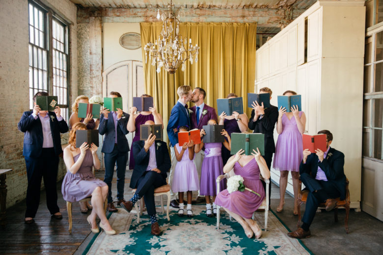 Wedding party in purple and navy holding books in front of faces as two grooms kiss under chandelier 