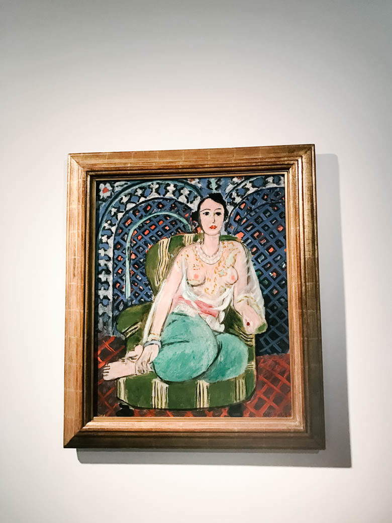 painting of dark haired woman in green chair wearing teal pants and a see-through blouse