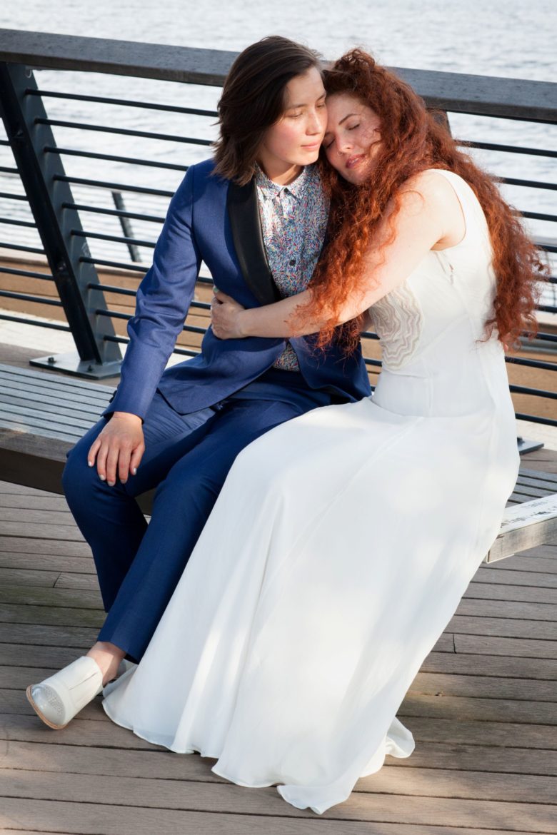 Couple in wedding attire embracing on bench near water