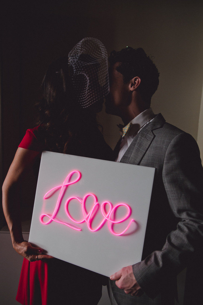 Couple kissing, holding pink on white EL wire sign that reads "Love"