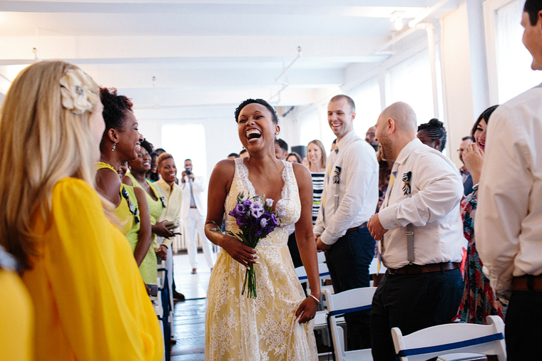 Bride laughing joyfully as she walks down the aisle in a bright white room holding a bouquet of purple flowers