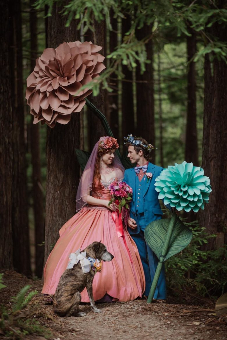 A bride wearing a dark pastel pink dress stands with her groom in a forest with giant towering paper flowers