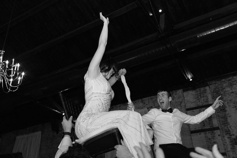 A bride & groom hold onto a napkin together as they are lifted up in chairs on the dance floor