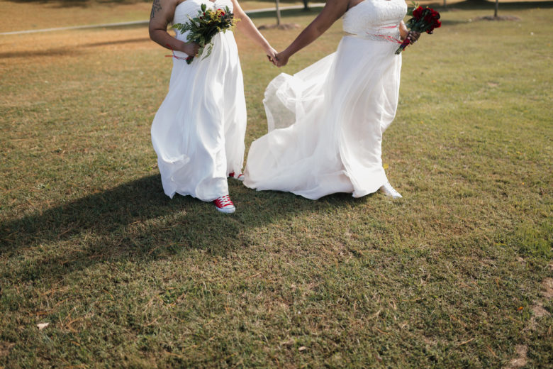 Two brides in a field wearing sneakers, holding hands and bouquets