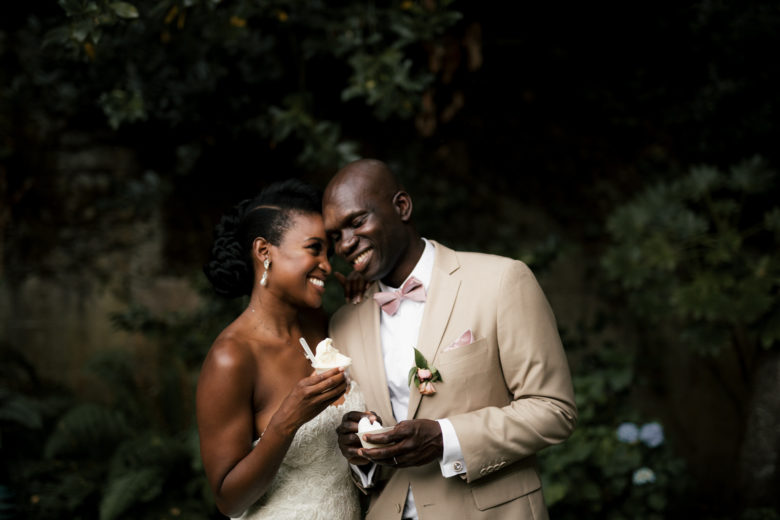 Bride and groom smiling, leaning into each other, as they hold sweet treats