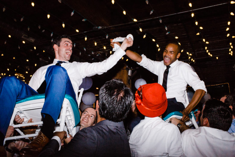 two joyful grooms being hoisted in chairs by wedding guests beneath illuminated string lights