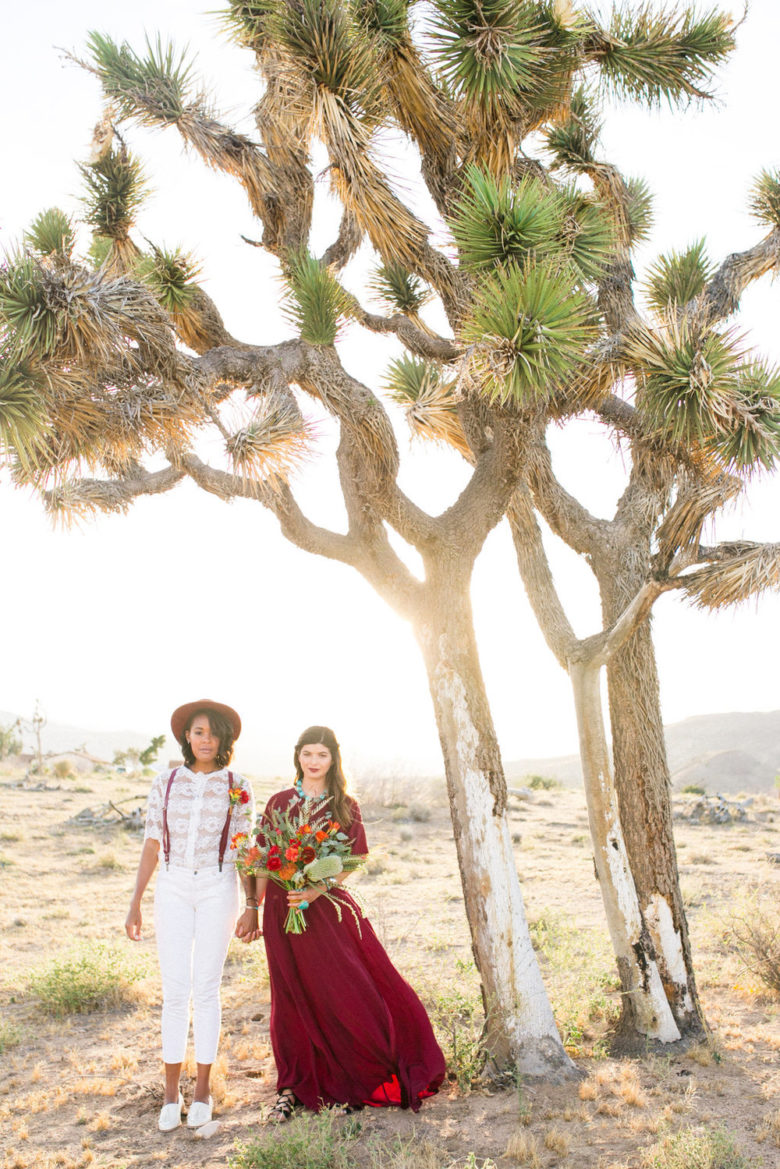 a married couple of women stand next to a giant joshua tree as the sun sets behind them