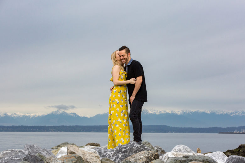 woman in yellow flower dress kissing man in black's cheek as he laughs, as they stand on rocks in front of mountains across water