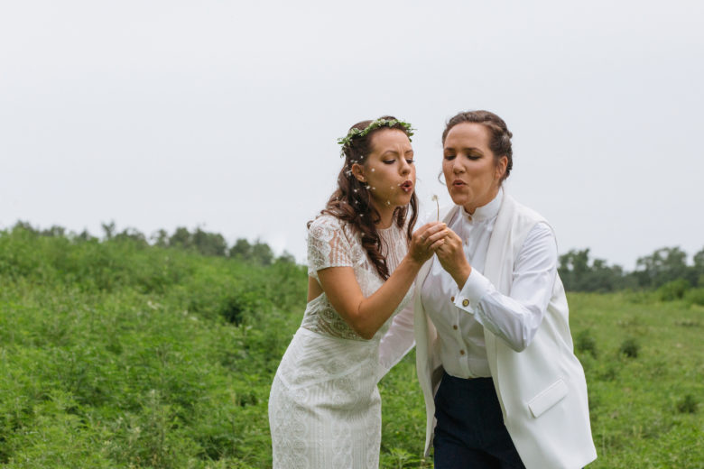 two brides in a field blowing on a dandelion 