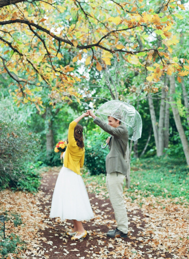 groom with clear umbrella twirling bride wearing yellow sweater under tree with yellow and orange trees in woods