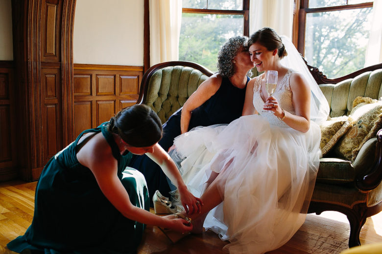 bride sitting on green settee, holding a glass of champagne, as her mother kisses her cheek and her bridesmaid fastens her shoe