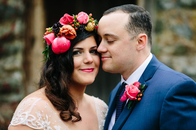 Bride with long dark hair and flower crown looking at you, smiling, with her face near the groom in a blue suit with pink flower boutonniere