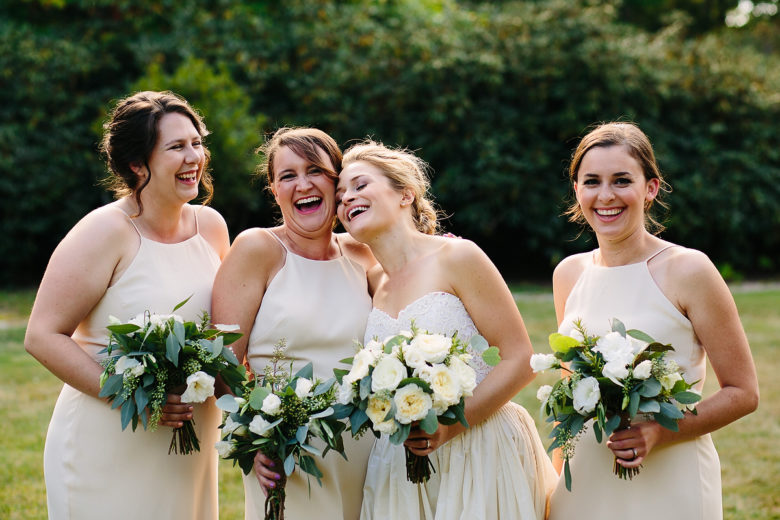 bride laughing with three bridesmaids in ivory dresses, all holding green and white bouquets