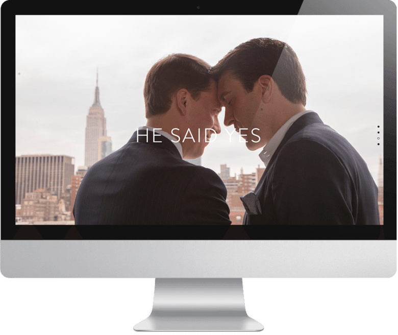 Two happy men in suits with their foreheads together overlooking a city skyline with text overlay that reads "he said yes" on a computer monitor