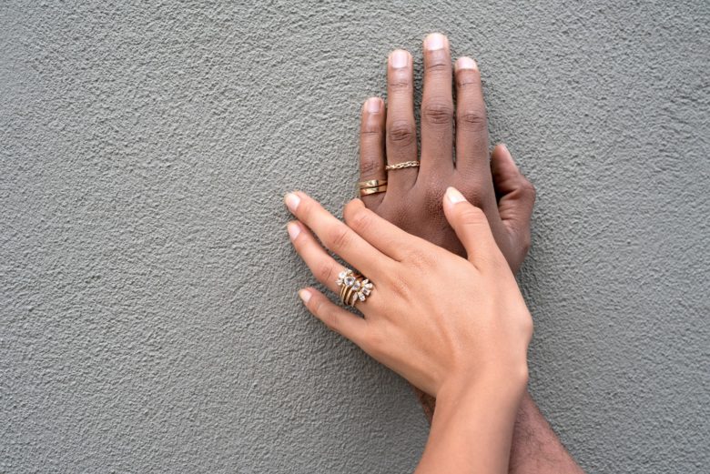 Caucasian hand with multiple diamond wedding rings on top of a black hand also wearing gold wedding rings against a dark gray wall. 