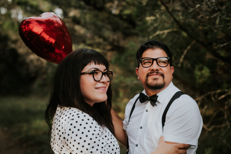couple wearing black and white, both with black frame glasses, looking off-camera, smiling, holding a red mylar balloon