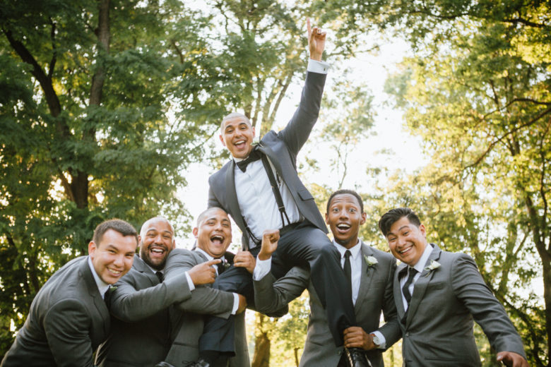 a group of jubilant men in suits hoisting groom up in a park
