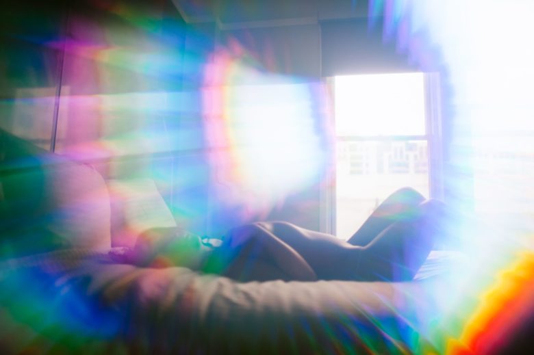 A lone person lies naked on a bed with the light streaming in from a window - lens flares obscuring the view