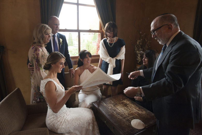 two brides signing ketubah with rabbi, surrounded by family
