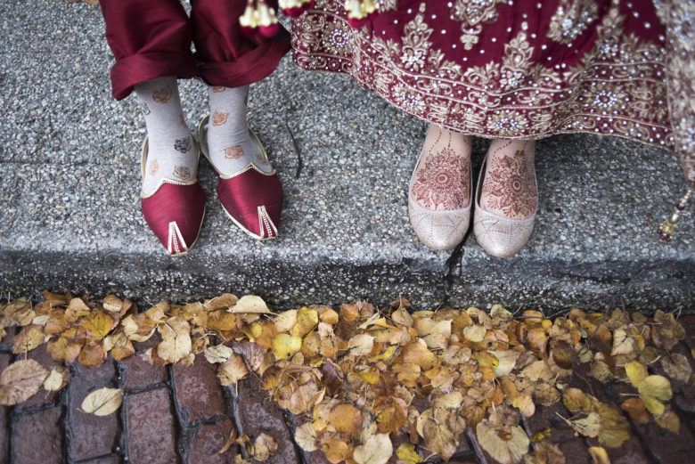 Two pairs of feet, one in colorful red slippers, the other in taupe flats with the top of the feet decorated in red henna, standing on a curb with yellow autumn leaves