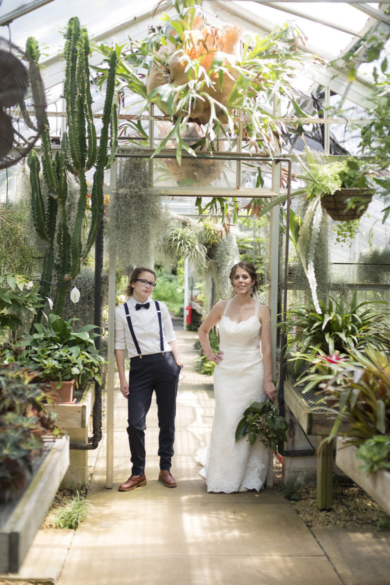 wedding couple, one in a white dress holding bouquet, the other in black pants with suspenders and a bow tie, leans in the doorway in a greenhouse 
