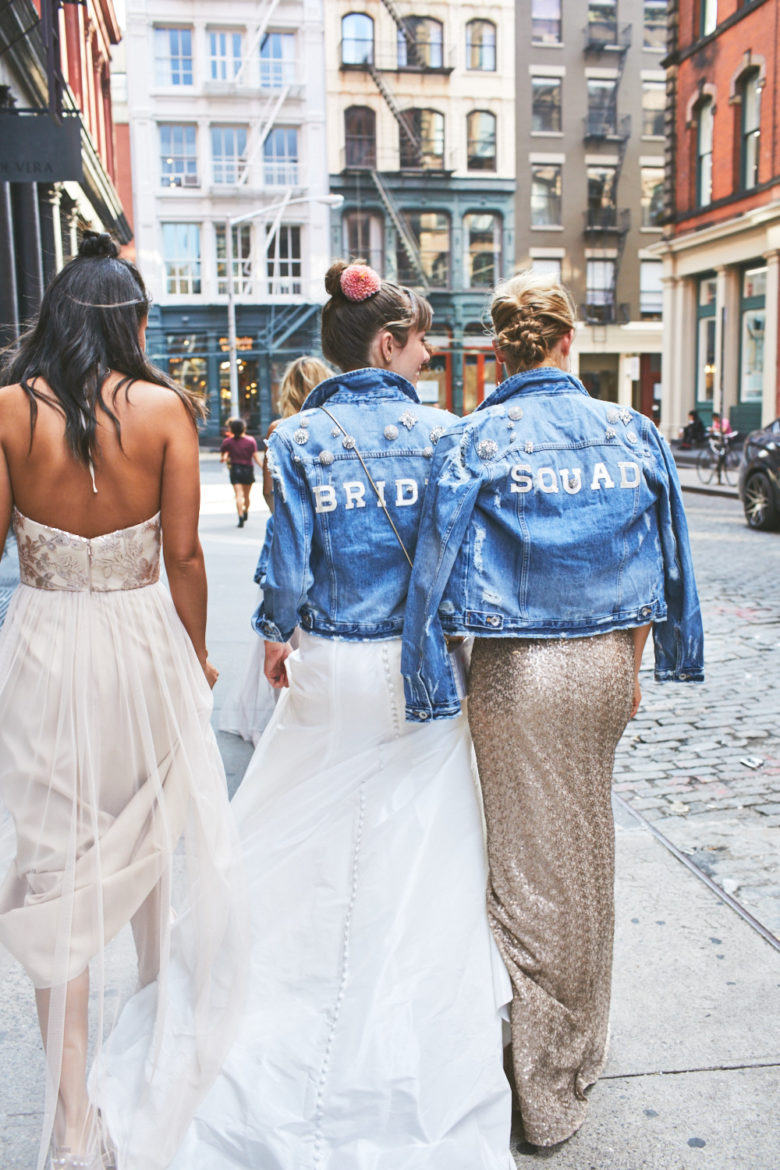 Bridesmaids and bride walking together on city streets wearing denim jackets over their dresses with the words BRIDE and SQUAD on the back.