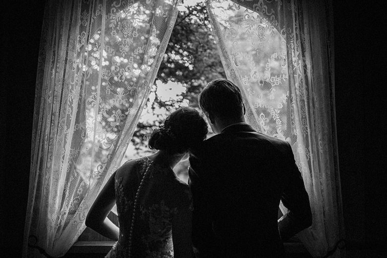 couple in wedding attire silhouetted in front of window with sheer embroidered curtains