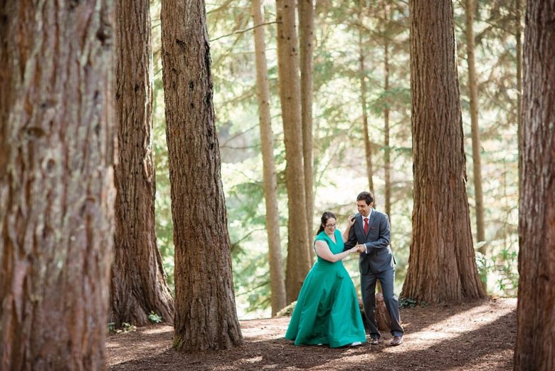 Woman in emerald green wedding dress holds hand, dancing with besuited man in a forest