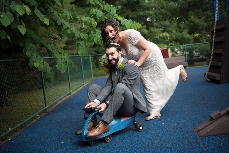 woman in wedding gown pushing a man in a suit and yarmukle riding a small sit-down scooter, both smiling and acting silly