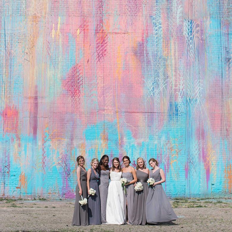 Bridal party in full-length grey dresses surround a bride in front of a pink, peach, blue, and purple watercolored wall