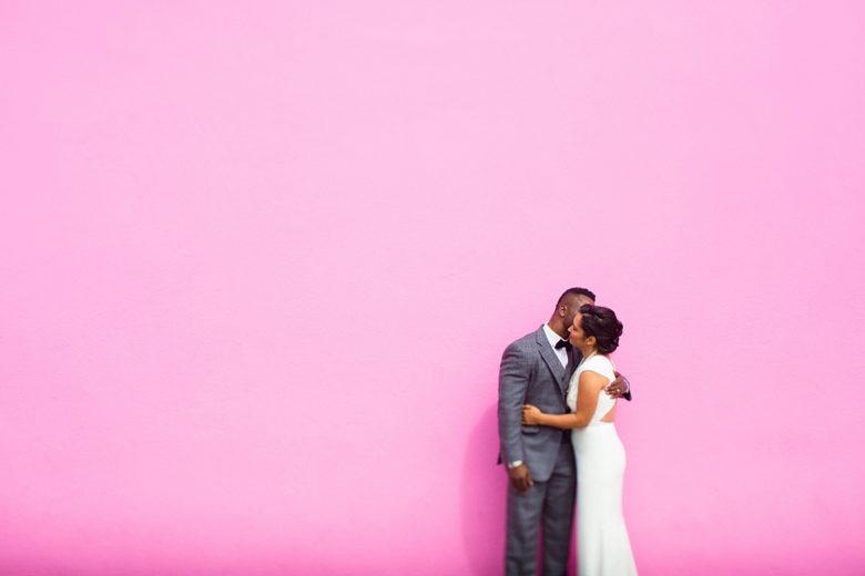 couple in wedding attire embracing in front of a bright pink wall