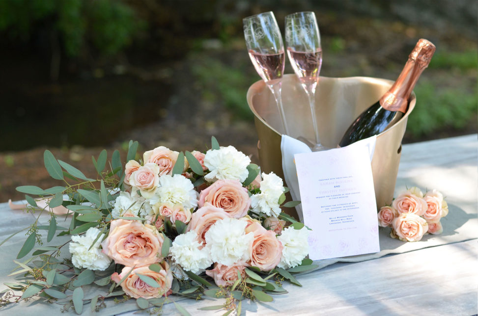 champagne glasses and bottle in gold holder on table with bouquet of peach roses and white carnations and eucalyptus
