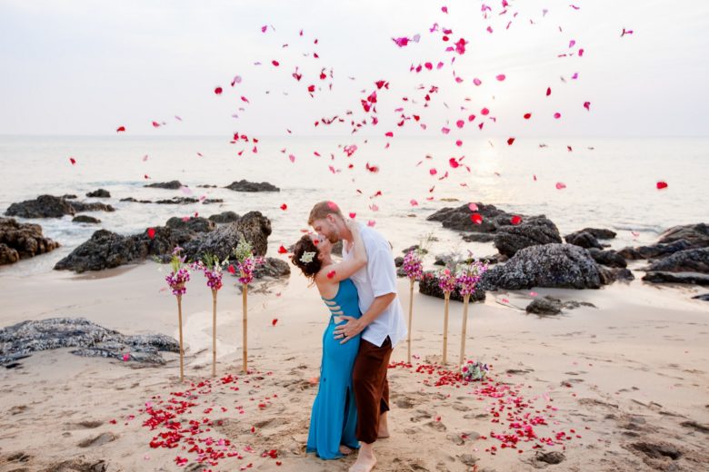 bride in a blue dress and groom kissing on a beach with red and pink flower confetti