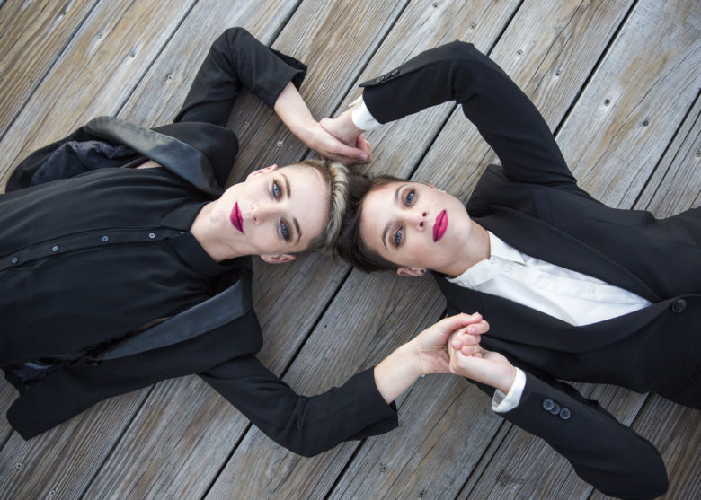 two women in black blazers and red lipstick lying down and looking up at camera and holding hands