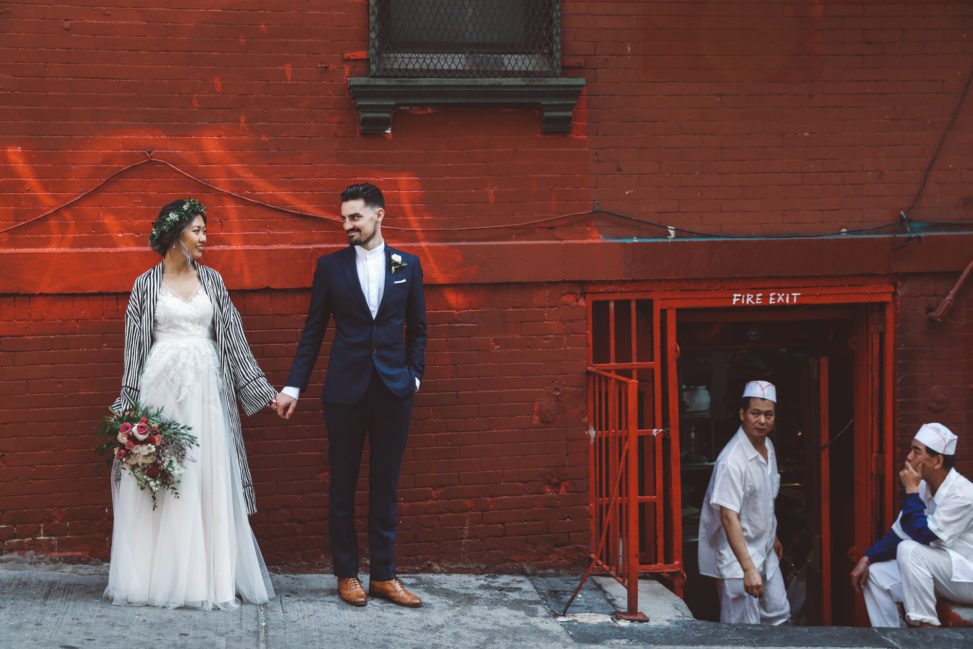 Couple in wedding finery holding hands, gazing at each other in front of a red brick wall as two restaurant workers take a cigarette break in an adjacent fire exit