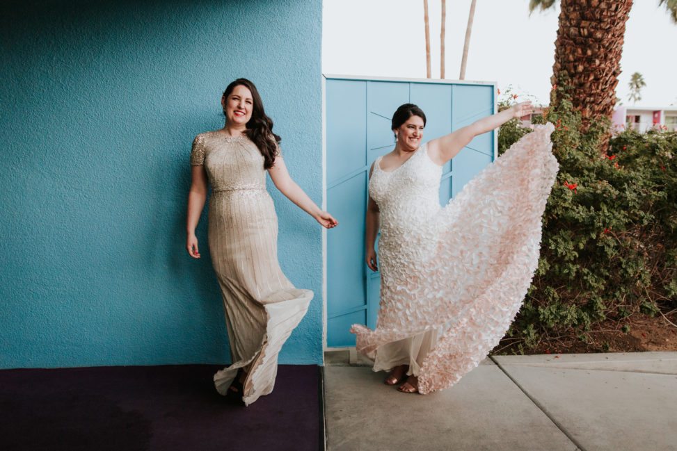 lesbian couple in wedding dresses standing in front of a blue wall at the Saguaro palm springs