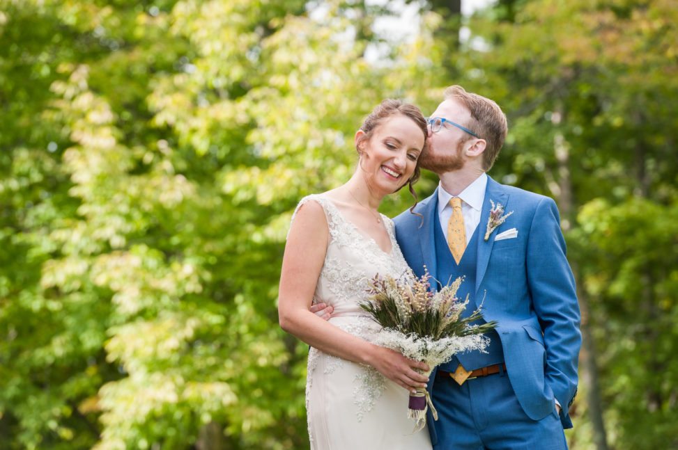 groom in blue suit with blue glasses kissing bride's temple in front of green trees