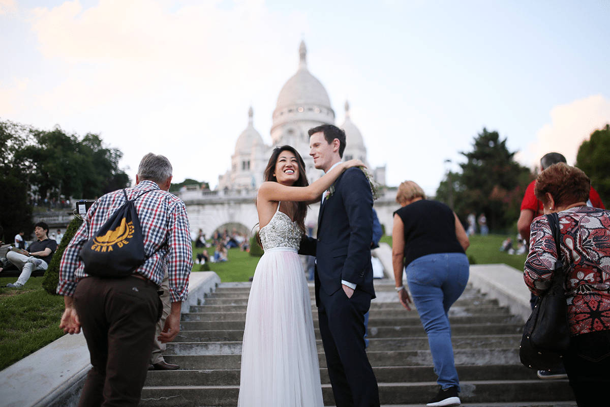A bride and groom smile at the bottom of the steps of Sacre Coeur as passerbys walk up the stairs toward the church.