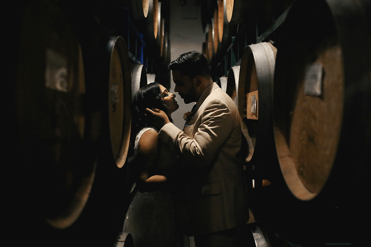 a man cradles a woman's head while standing in between rows of large wooden wine barrels