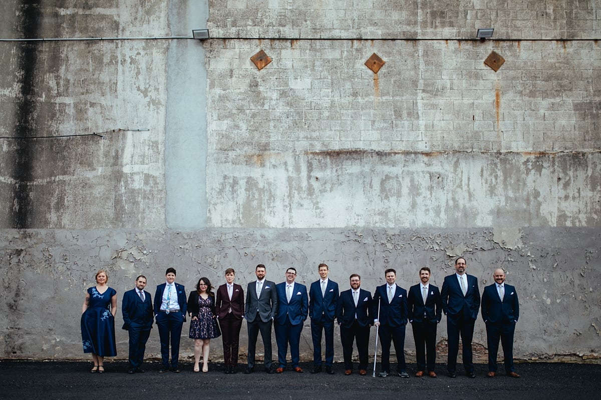 a wedding party lines up shoulder to shoulder and stands in front of a giant wall