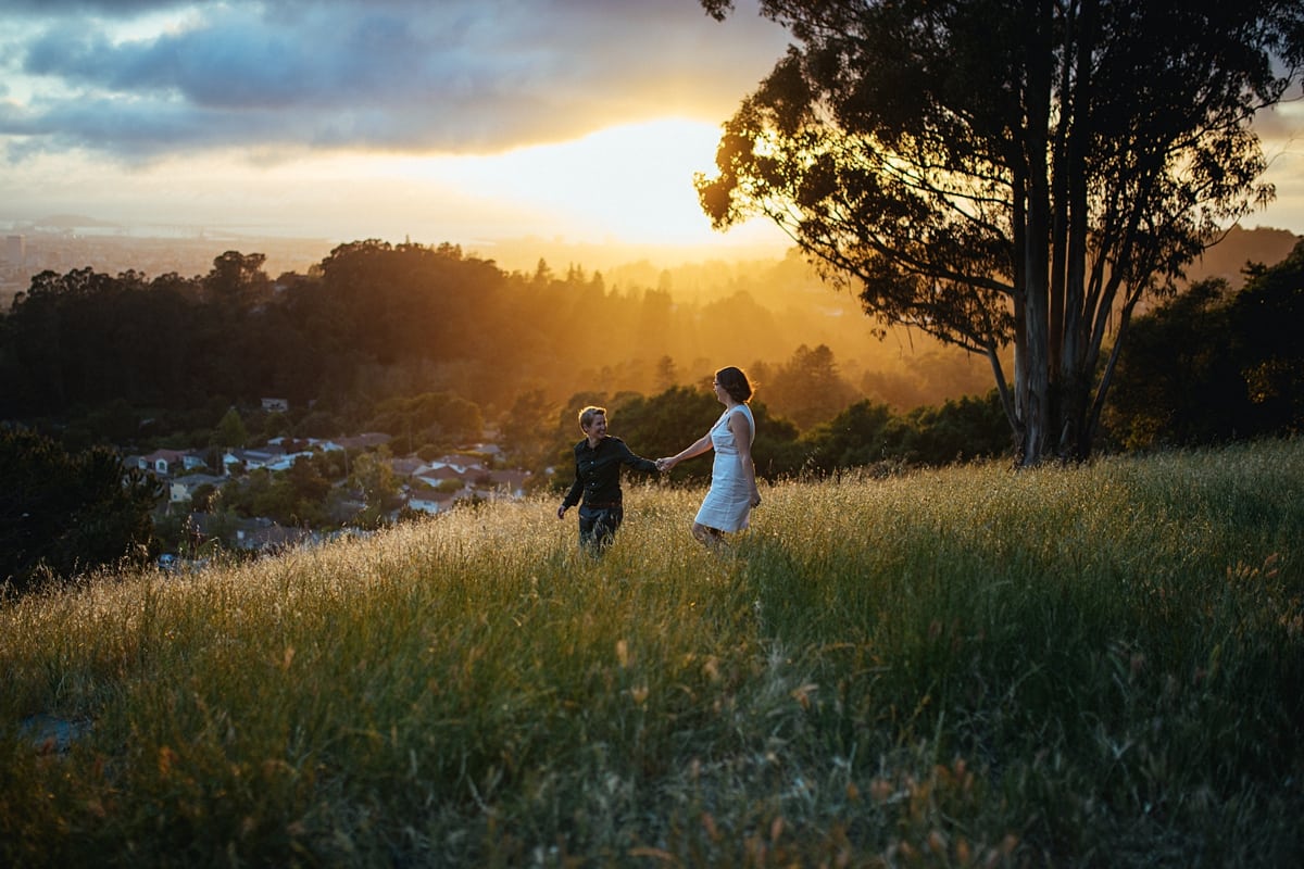 two persons hold hands and walk down a hill with a brilliant sunset showing the heavens behind them
